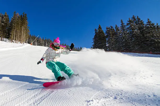 Žena na snowboardu sjíždějící zasněžený svah s prudkým obloukem, obklopená zimní krajinou s modrou oblohou a stromy v pozadí.