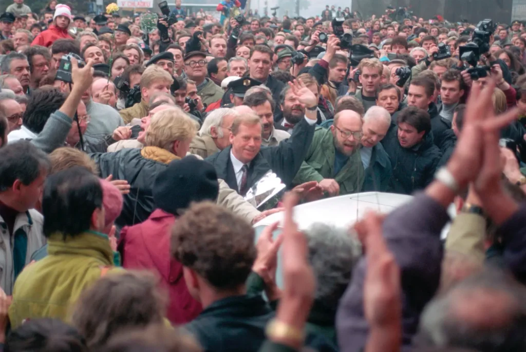 Václav Havel zdraví dav příznivců během sametové revoluce v roce 1990, obklopen lidmi oslavujícími naději a svobodu v Československu.