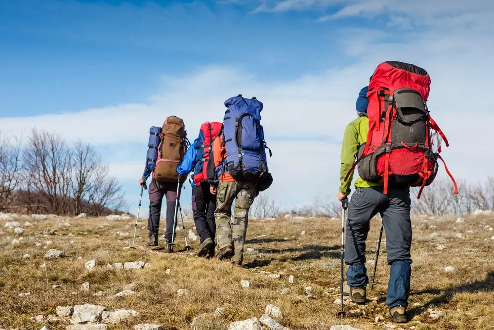 Skupina turistů s velkými batohy a trekingovými holemi stoupající po horské stezce během výšlapu, obklopeni přírodou a pod širým nebem.