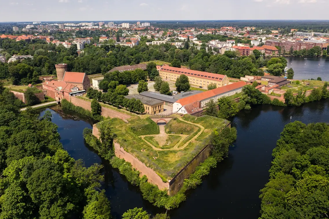 Letecký pohled na hrad Zitadelle Spandau v Berlíně, obklopený vodními kanály a zelení, s rozsáhlým opevněním a historickými budovami uprostřed městské krajiny.