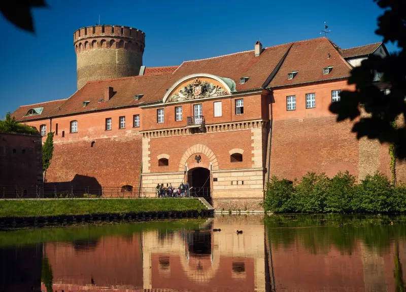 Hrad Zitadelle Spandau v Berlíně, zachycený za slunečného dne s klidnou vodní hladinou v popředí, odrážející masivní hradby a vstupní bránu s věží v pozadí.