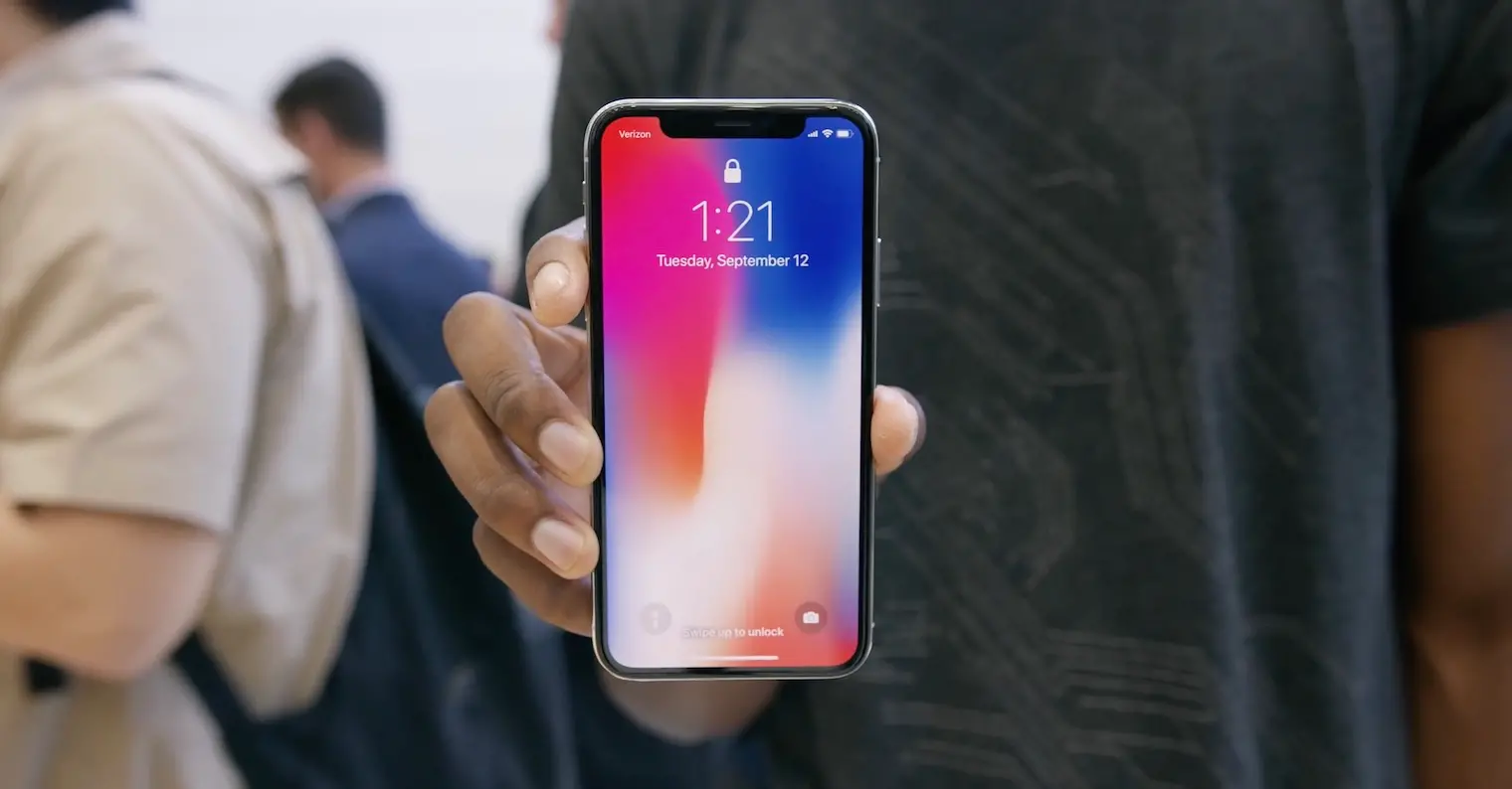 Uživatel drží iPhone X, který poprvé přinesl bezrámečkový design a Face ID. Na displeji je vidět zamčená obrazovka s charakteristickým výřezem (notch) pro senzory a kameru.