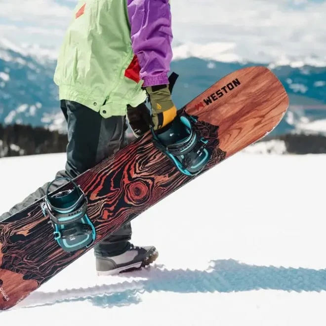 Snowboardista držící snowboard s dřevěným designem značky Weston, stojící na zasněženém svahu s výhledem na hory, ideální pro freeride i freestyle.