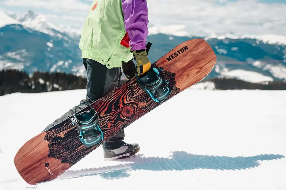 Snowboardista držící snowboard s dřevěným designem značky Weston, stojící na zasněženém svahu s výhledem na hory, ideální pro freeride i freestyle.