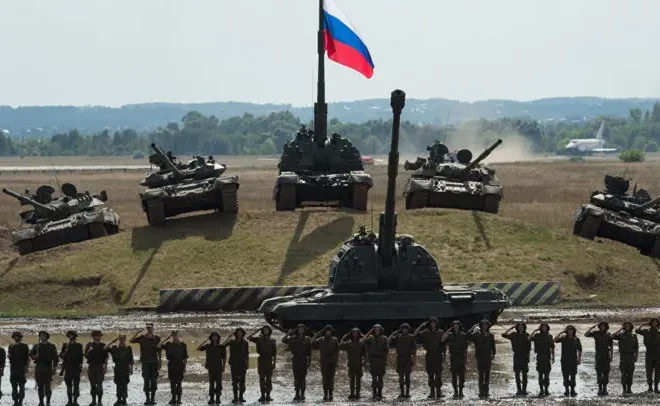 Ruská vojenská přehlídka s tanky a vojáky stojícími v řadě pod vlajkou Ruska na venkovském pozadí.