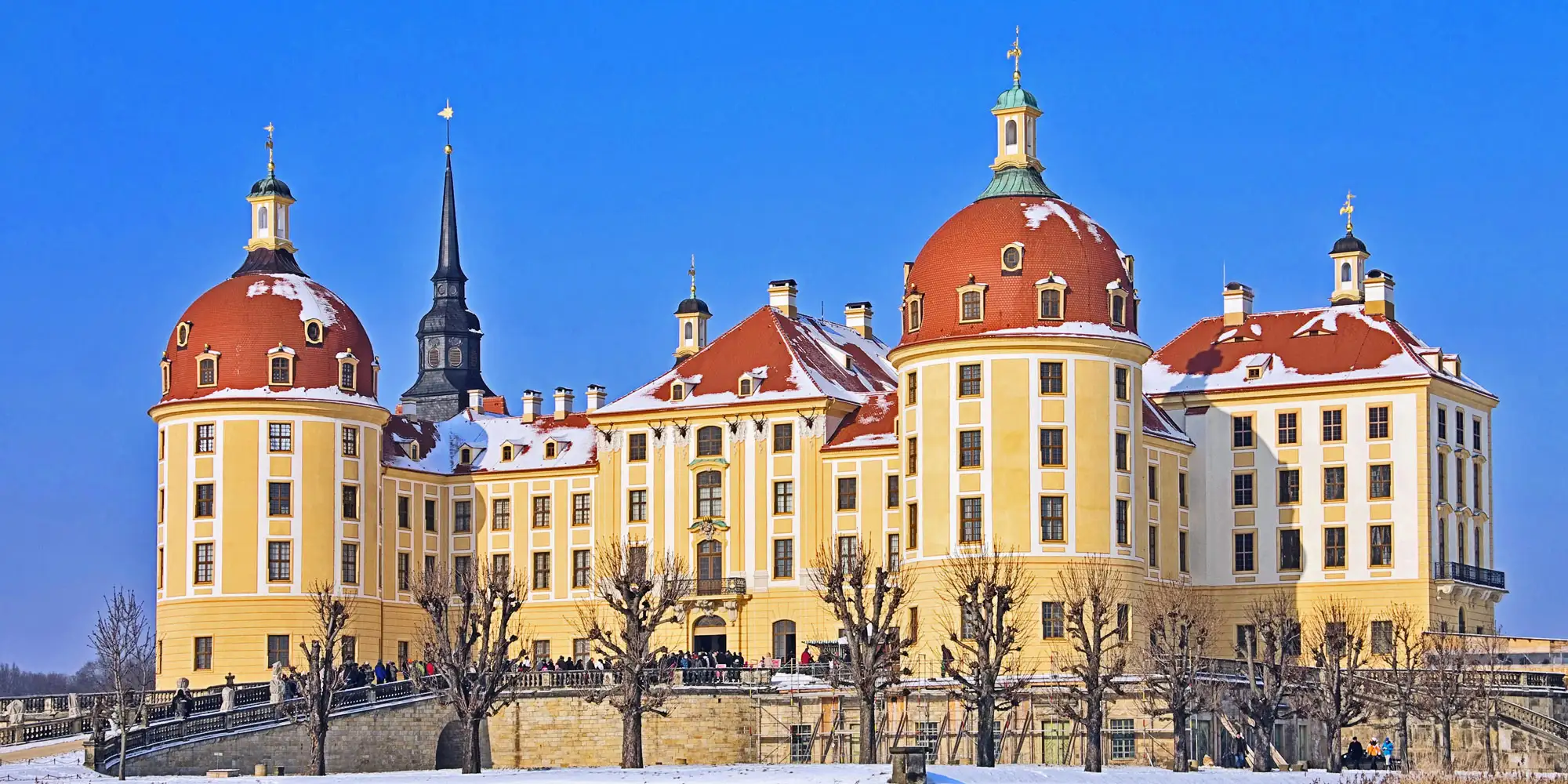 Zimní pohled na pohádkový zámek Moritzburg, známý z filmu „Tři oříšky pro Popelku“, s červenými střechami pokrytými sněhem, pod modrou oblohou.