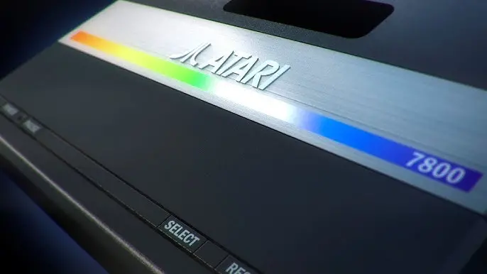 Detailní záběr na konzoli Atari 7800 s logem Atari a barevným pruhem na horní straně, tlačítky „Select“ a „Reset“ v popředí.