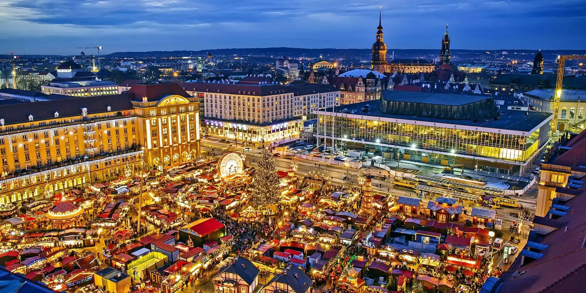 Panoramatický pohled na vánoční trhy v Drážďanech s krásně ozdobenými stánky, historickými budovami a večerním osvětlením.