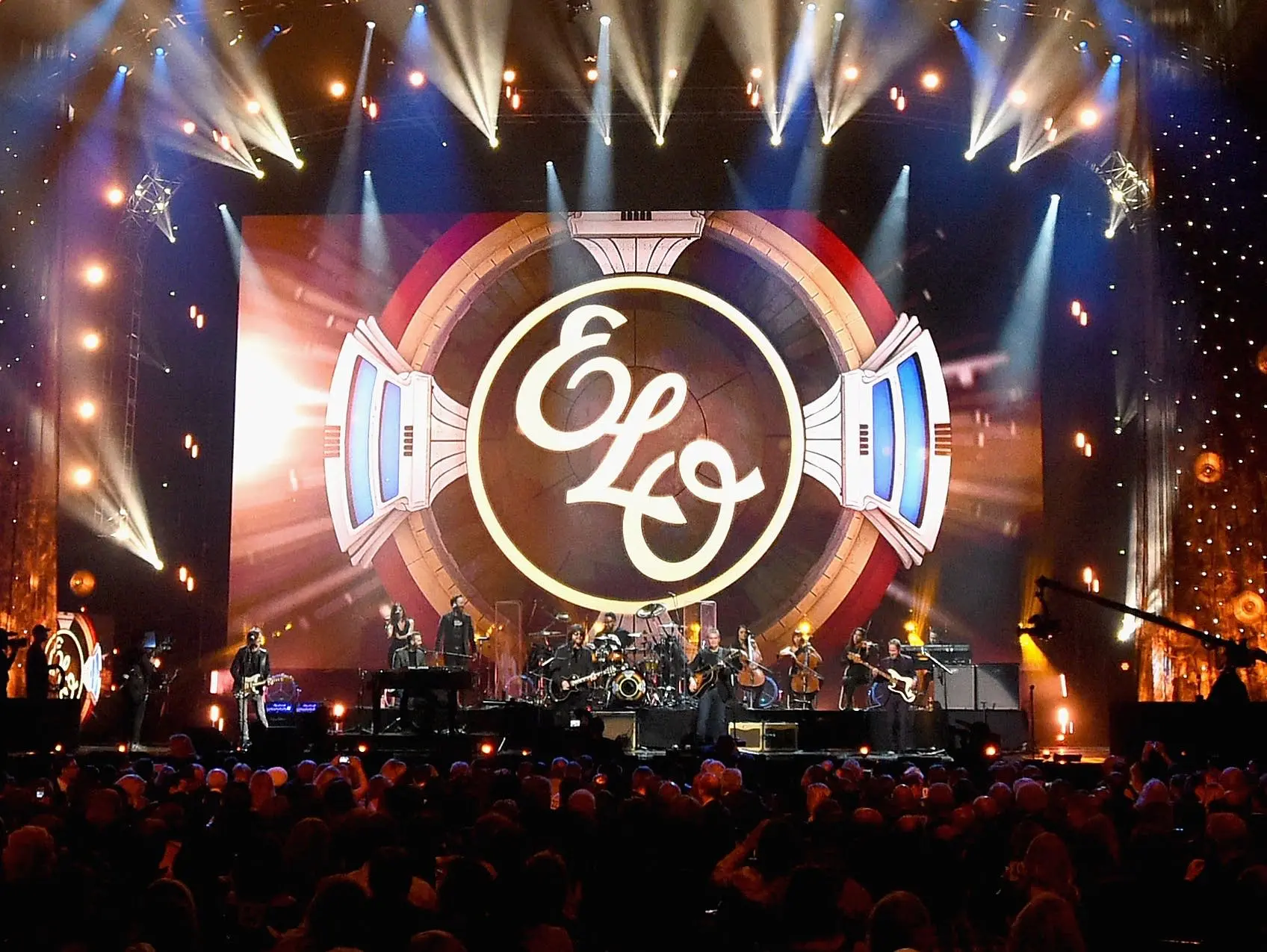 Electric Light Orchestra na pódiu během koncertu, velké logo ELO na pozadí a světelná show zachycující atmosféru vystoupení.