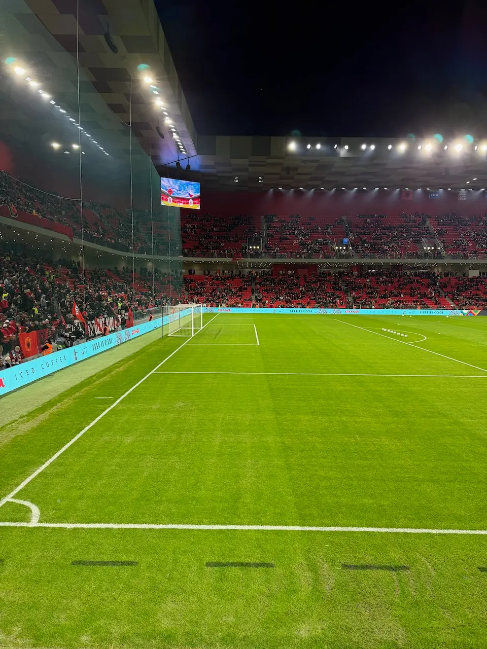 Fotbalové hřiště na Air Albania Stadionu v Tiraně během zápasu, zobrazující dokonale upravený trávník, osvětlené tribuny plné fanoušků a moderní stadionové zázemí, které vytváří atmosféru vrcholového sportovního zážitku.