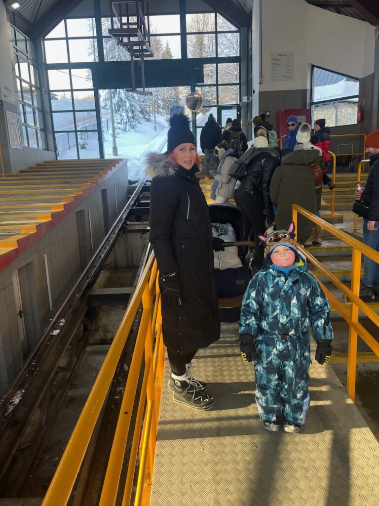 Matka s dítětem čekající na lanovku Gubałówka v interiéru stanice, se zasněženými stromy viditelnými za velkými okny.