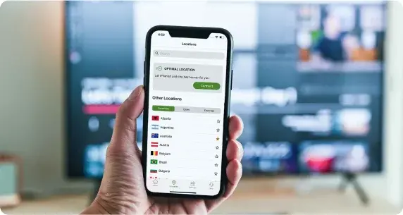 Mobilní telefon s otevřenou aplikací IPVanish VPN, kde uživatel vybírá optimální lokaci serveru. Na obrazovce je seznam zemí, jako Albánie, Austrálie a Rakousko, a tlačítko pro připojení k serveru. V pozadí televize s přehrávaným obsahem.