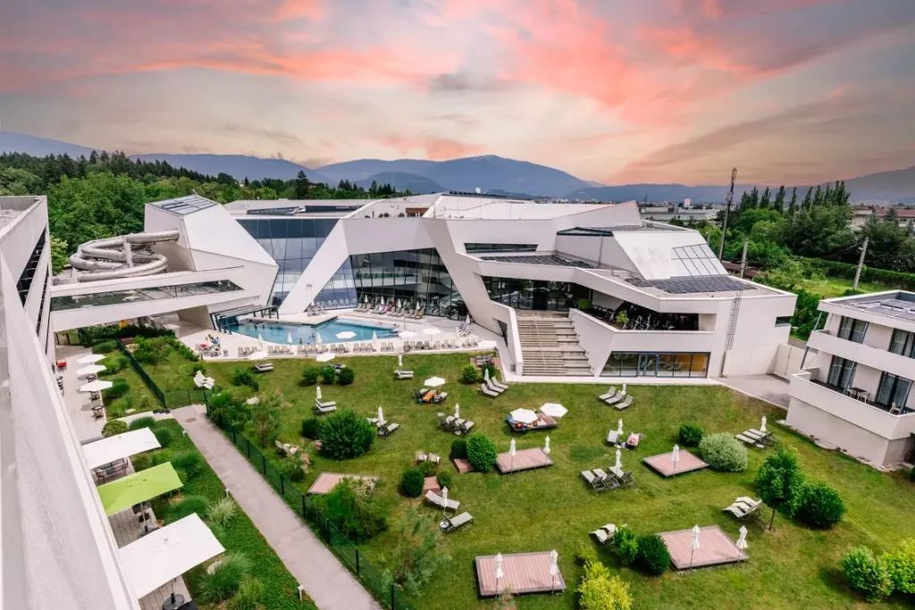 Exteriér termálních lázní Kärnten Therme v rakouských Korutanech při západu slunce. Moderní architektura s venkovním bazénem a zelenými relaxačními plochami v prostředí alpské přírody poskytuje dokonalý odpočinek.