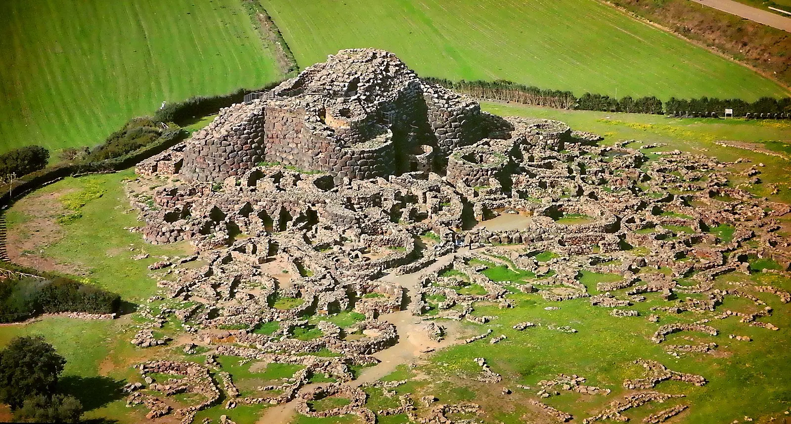 Letecký pohled na archeologické naleziště Nuraghe Su Nuraxi na Sardinii, obklopené zelenými poli. Kamenná struktura z doby bronzové s kruhovými stavbami ukazuje unikátní architekturu starověké nuragské civilizace.