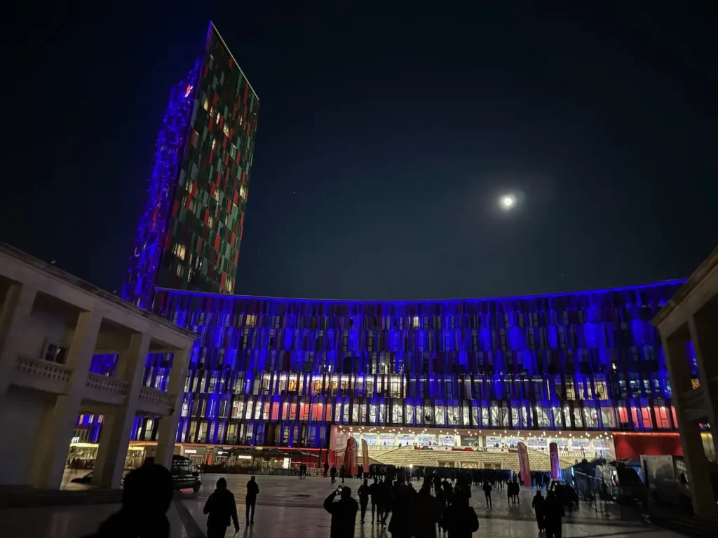 Osvětlený Air Albania Stadion v nočním záběru, zobrazující ikonickou věž a moderní fasádu stadionu s modrým a červeným podsvícením, pod jasnou noční oblohou s úplňkem, v popředí siluety návštěvníků.