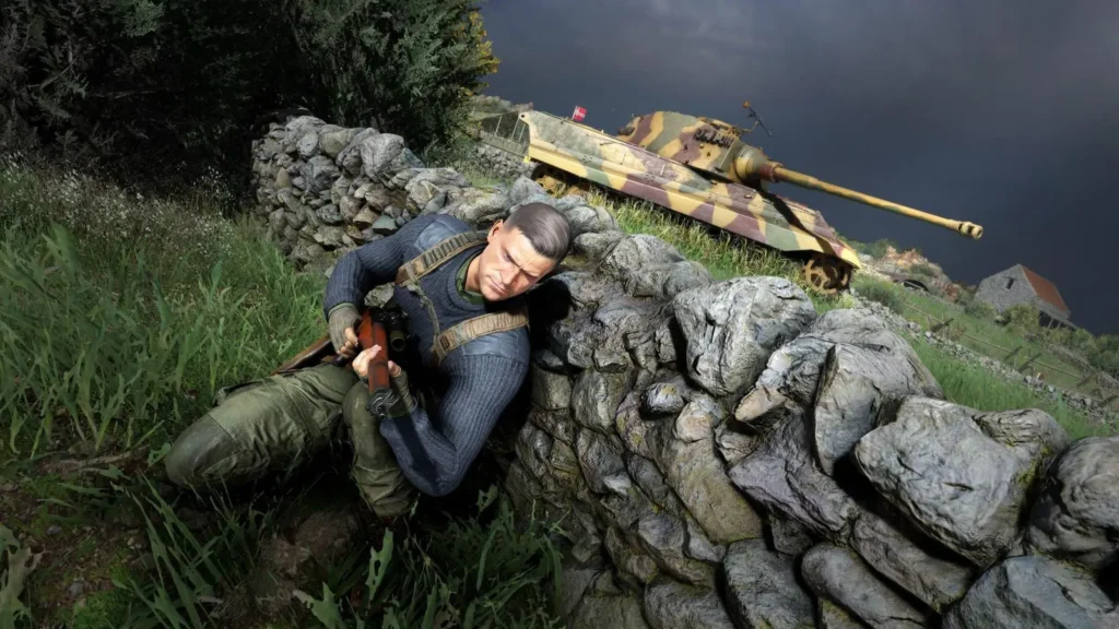 Karl Fairburne, hlavní postava Sniper Elite 5, se kryje za kamenitou zídkou na venkově, zatímco v těsné blízkosti za ním stojí maskovaný německý tank. Hrdina sleduje okolí s připravenou puškou, zatímco temné nebe přidává scéně dramatický nádech.