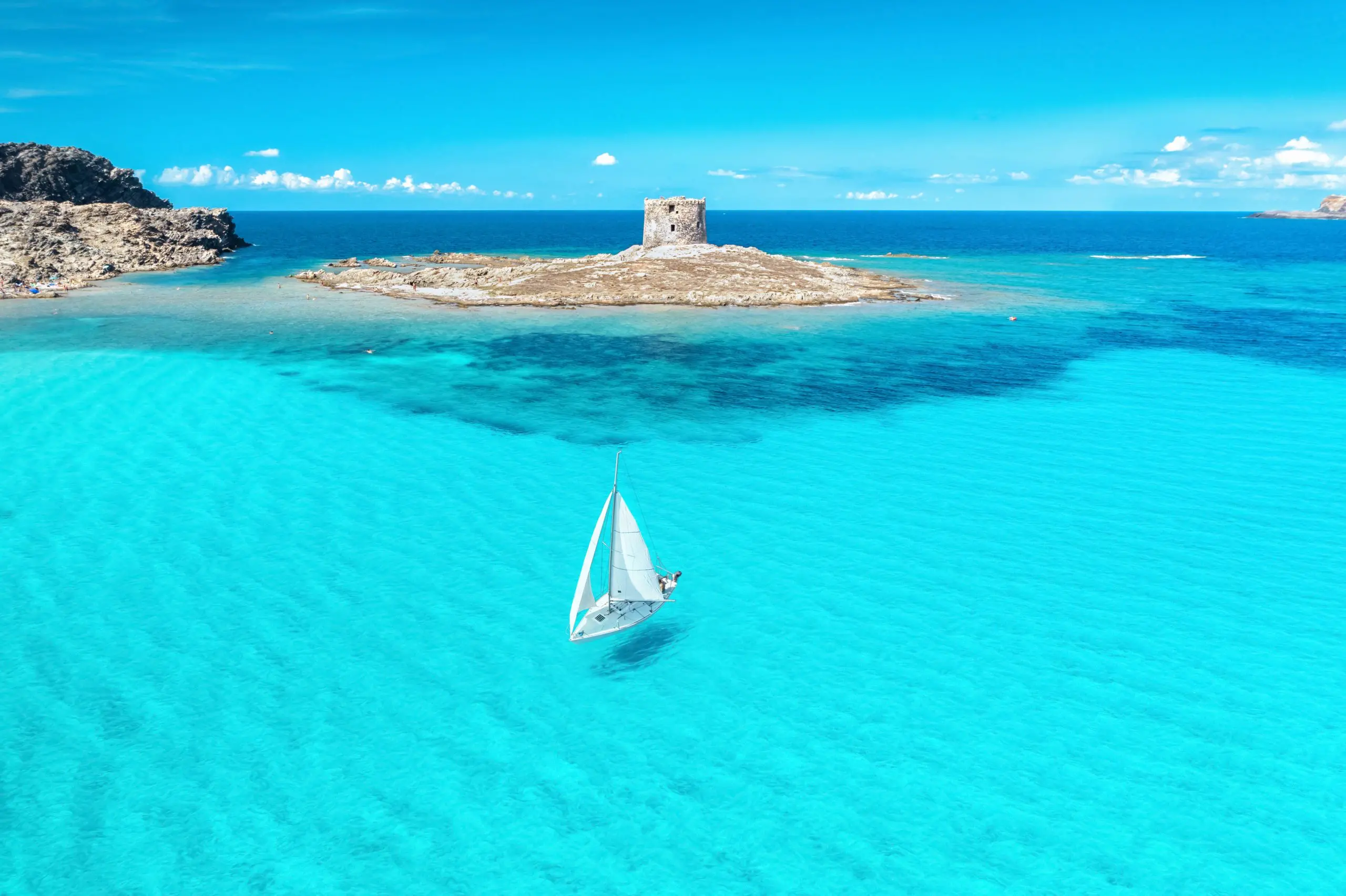 Křišťálově čistá tyrkysová voda u pláže Spiaggia della Pelosa na Sardinii s malým ostrůvkem a historickou věží Torre della Pelosa v pozadí, doplněná plachetnicí na klidné hladině moře.