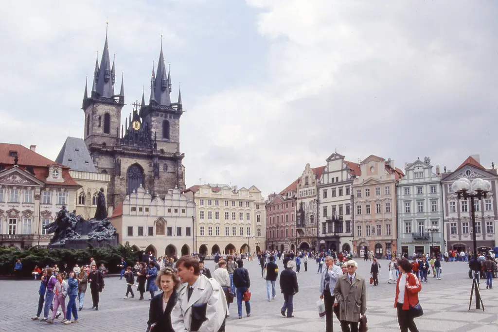 Staroměstské náměstí v Praze v roce 1991. Na fotografii je vidět Týnský chrám a socha Jana Husa obklopená lidmi. V pozadí se tyčí historické domy s pestrými fasádami, zatímco na náměstí se pohybují lidé v typickém oblečení počátku 90. let, což zachycuje atmosféru doby krátce po Sametové revoluci.