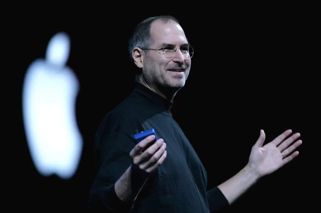 Steve Jobs, spoluzakladatel společnosti Apple, přednáší na pódiu v černém roláku s logem Apple v pozadí.