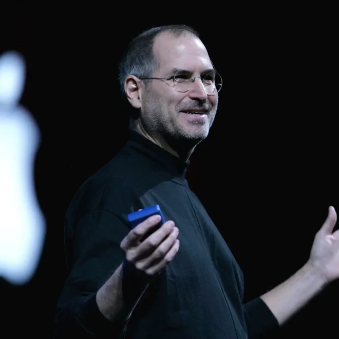 Steve Jobs, spoluzakladatel společnosti Apple, přednáší na pódiu v černém roláku s logem Apple v pozadí.