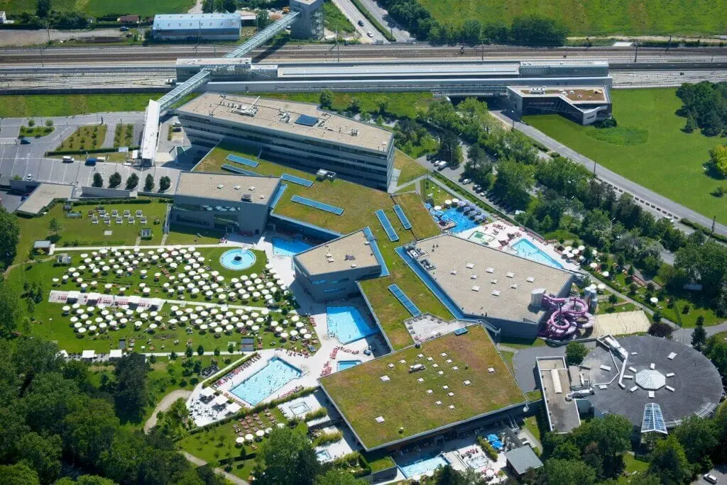 Letecký pohled na moderní termální lázně Therme Wien ve Vídni, s rozsáhlým komplexem bazénů, zelenými střechami a venkovními plochami pro relaxaci. Výjimečné místo pro odpočinek uprostřed městského prostředí.