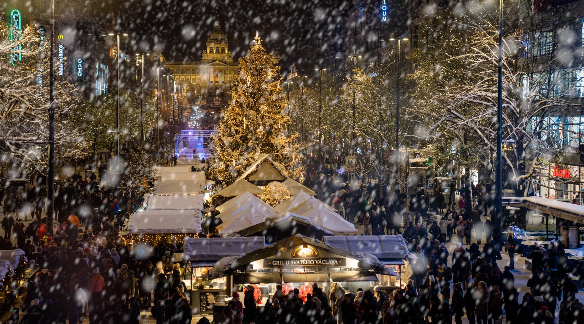 Vánoční strom a adventní trhy na Václavském náměstí v Praze, pokryté sněhem a obklopené lidmi v kouzelné atmosféře zimního večera.