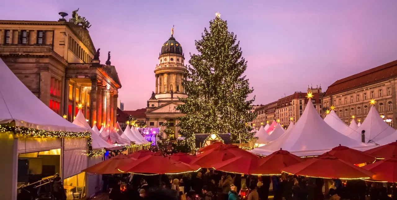 Pohled na vánoční trhy na náměstí Gendarmenmarkt v Berlíně, obklopené historickými budovami s vánoční výzdobou a velkým osvětleným stromem.