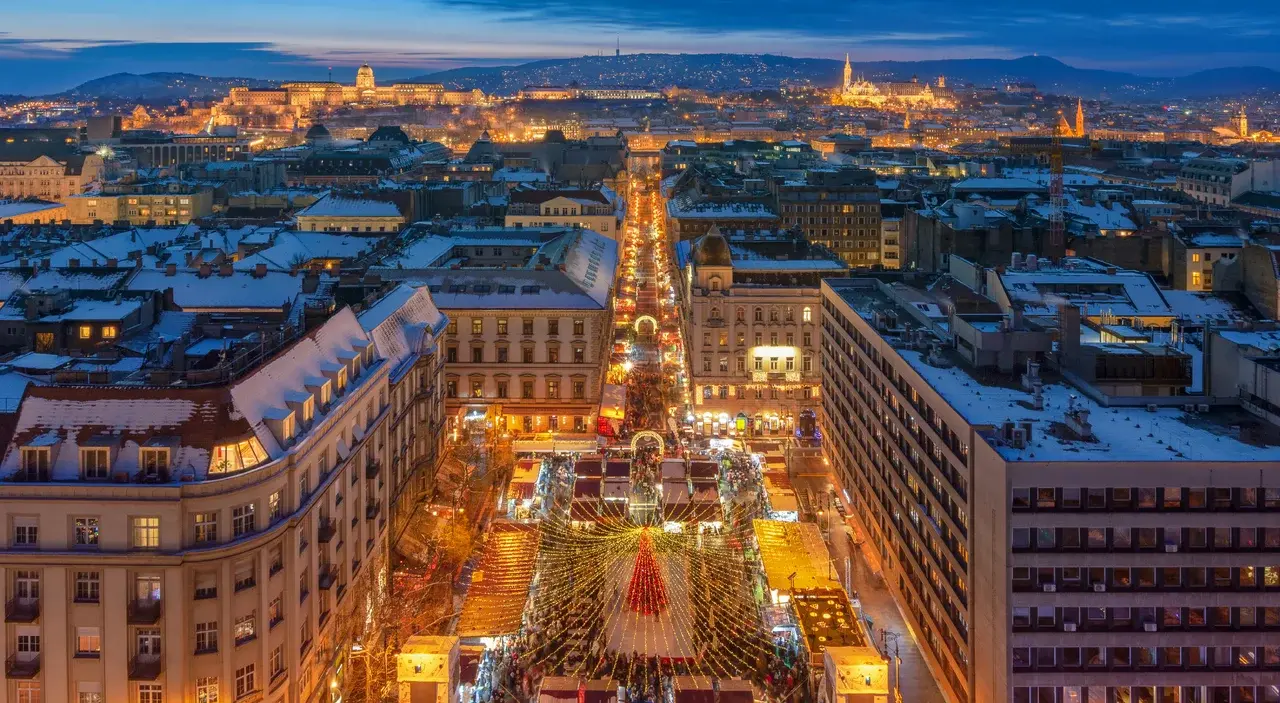 Nádherný pohled na vánoční trhy v Budapešti na náměstí Vörösmarty s osvětlenými stánky a výhledem na slavné památky ve večerní atmosféře.