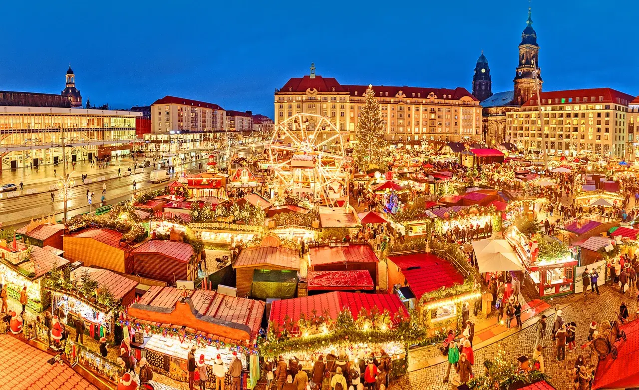 Pohled na vánoční trhy Štrýclmarkt v Drážďanech, plné barevně osvětlených stánků a velkého ruského kola v srdci města.