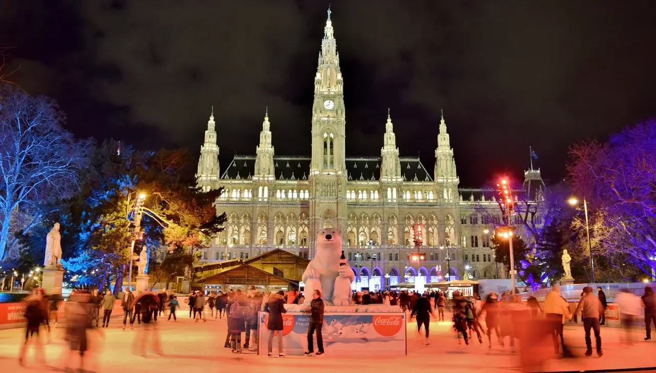 Pohled na vánoční trhy na Radničním náměstí ve Vídni, s ledovým kluzištěm a vánočními stánky v krásně nasvíceném prostoru před radnicí.