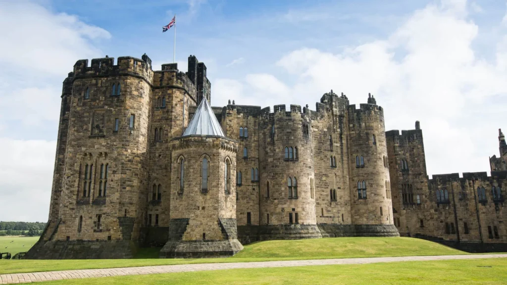 Historický hrad Alnwick v Anglii s typickou kamennou architekturou, věžemi a britskou vlajkou na pozadí modré oblohy.