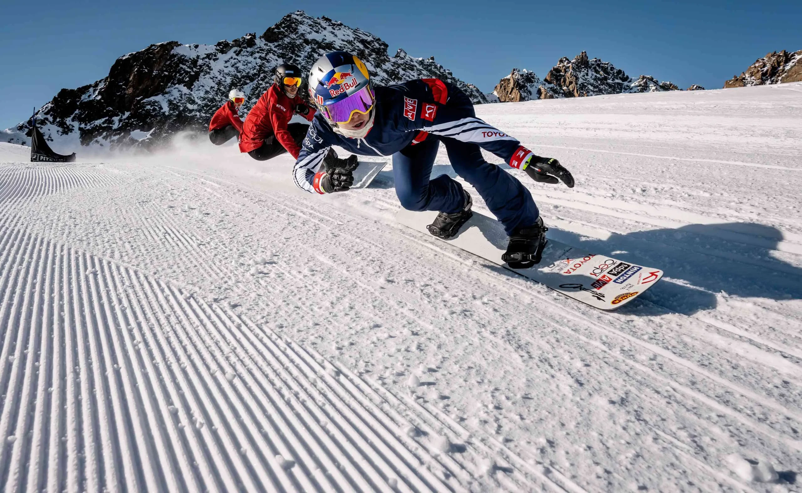 Eva Adamczyková závodí na snowboardu v Rakousku, na upravené sjezdovce, v závodní poloze s logem Red Bull na helmě.