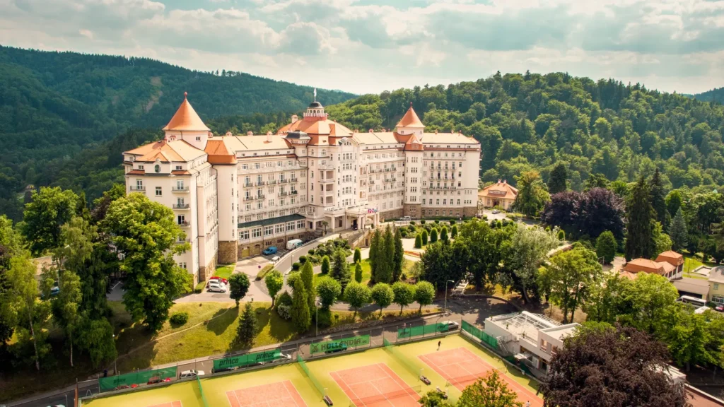 Hotel Imperial Karlovy Vary obklopený zelení s výhledem na tenisové kurty a okolní lesy za slunečného dne.