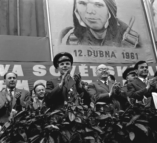 Jurij Gagarin stojí na pódiu v uniformě, obklopený československými a sovětskými představiteli, před plakátem oslavujícím jeho historický let 12. dubna 1961. Fotografie zachycuje jeho triumfální přijetí během návštěvy Československa, kde byl uctíván jako první člověk ve vesmíru.