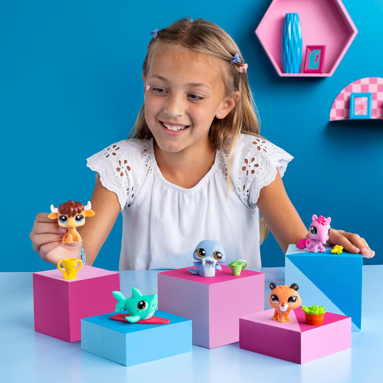 Usměvavá holčička si hraje s figurkami Littlest Pet Shop, které jsou vystavené na barevných růžových a modrých podstavcích. Figurky zahrnují různá zvířátka, jako je býček, delfín, liška a další, na pozadí s modrou stěnou a dekoracemi.