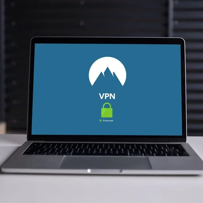 Otevřený notebook na stole se zobrazenou VPN aplikací, která ukazuje aktivní zabezpečení připojení prostřednictvím zelené ikony zámku. Pozadí je rozostřené s tmavými žaluzie.