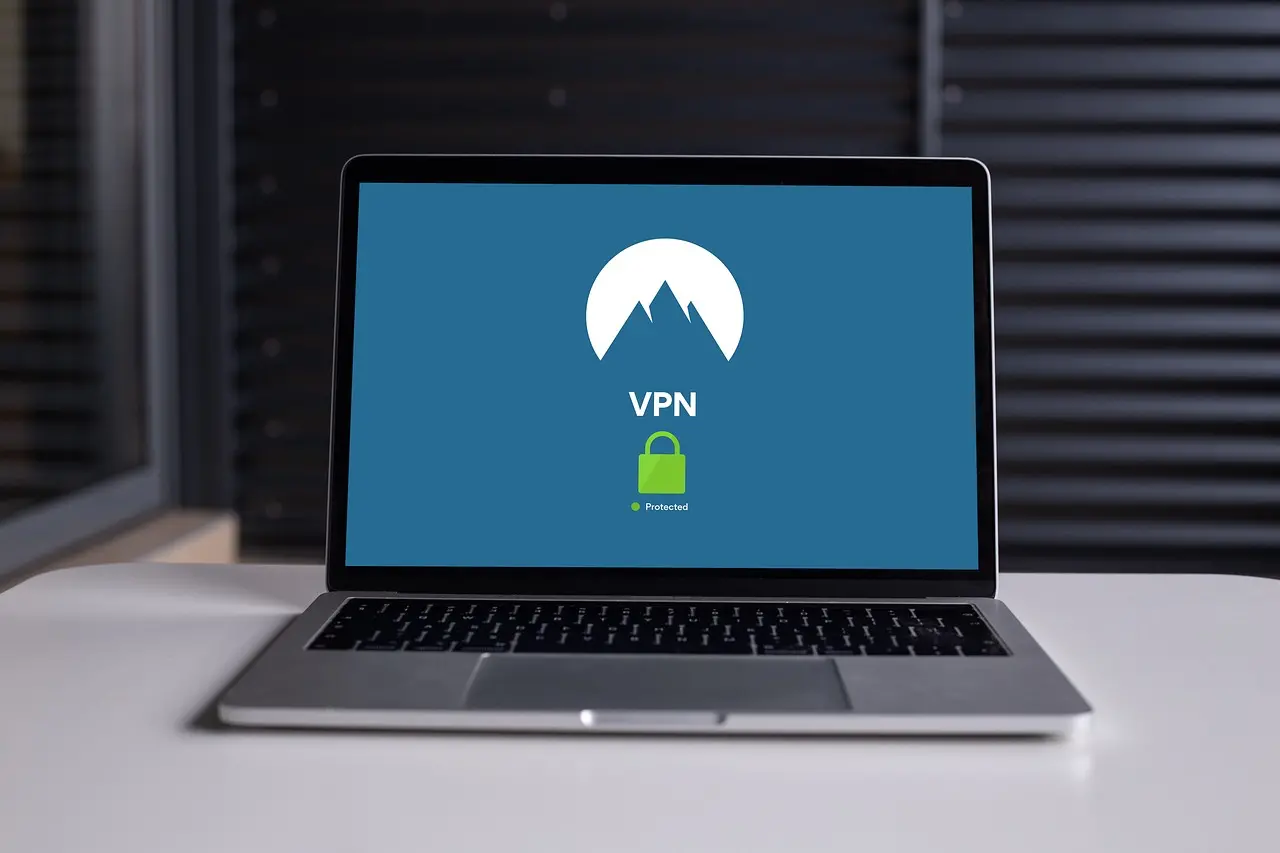 Otevřený notebook na stole se zobrazenou VPN aplikací, která ukazuje aktivní zabezpečení připojení prostřednictvím zelené ikony zámku. Pozadí je rozostřené s tmavými žaluzie.