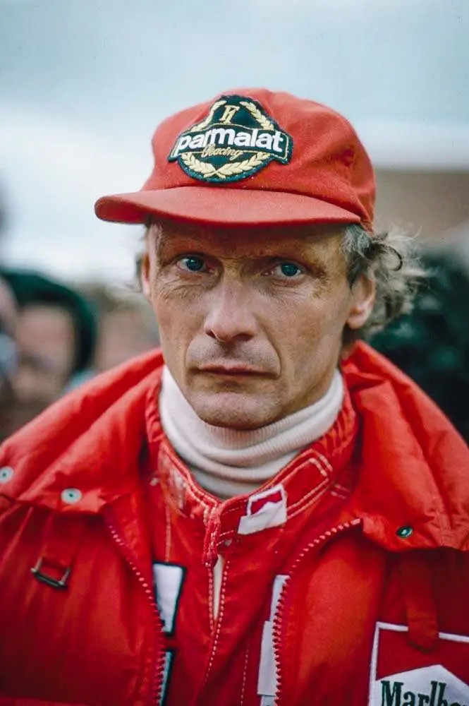 Niki Lauda v červené bundě Parmalat, s vážným pohledem během závodního dne, symbolizující jeho soustředění a profesionalitu.