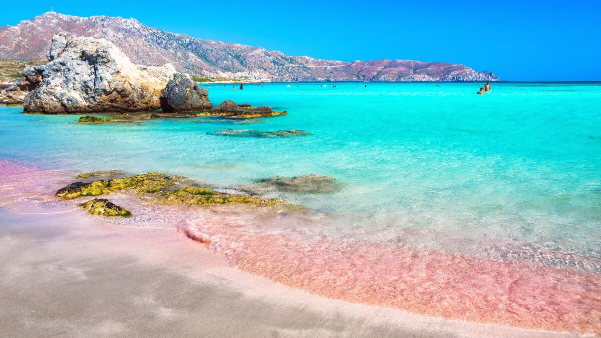 Pohled na růžový písek pláže Pink Sands Beach na Harbour Islandu, Bahamy, s průzračně modrou vodou a skalními útvary.