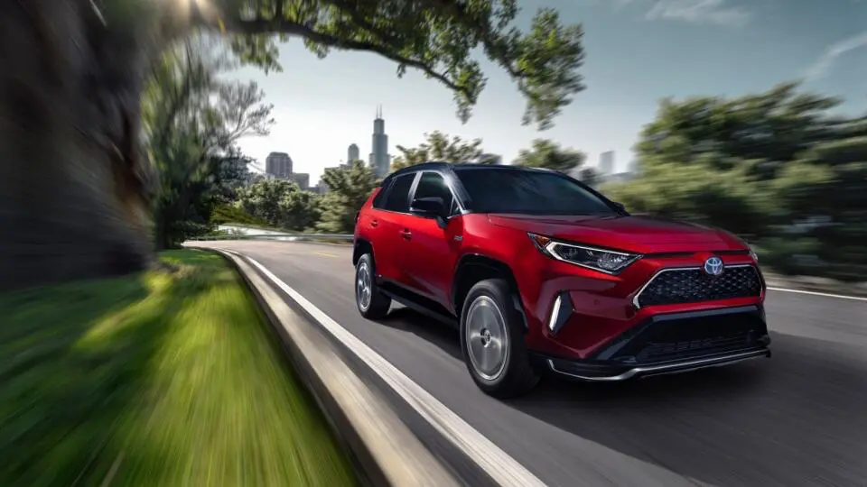 Červená Toyota RAV4 projíždí přírodní krajinou, v pozadí městské panorama.