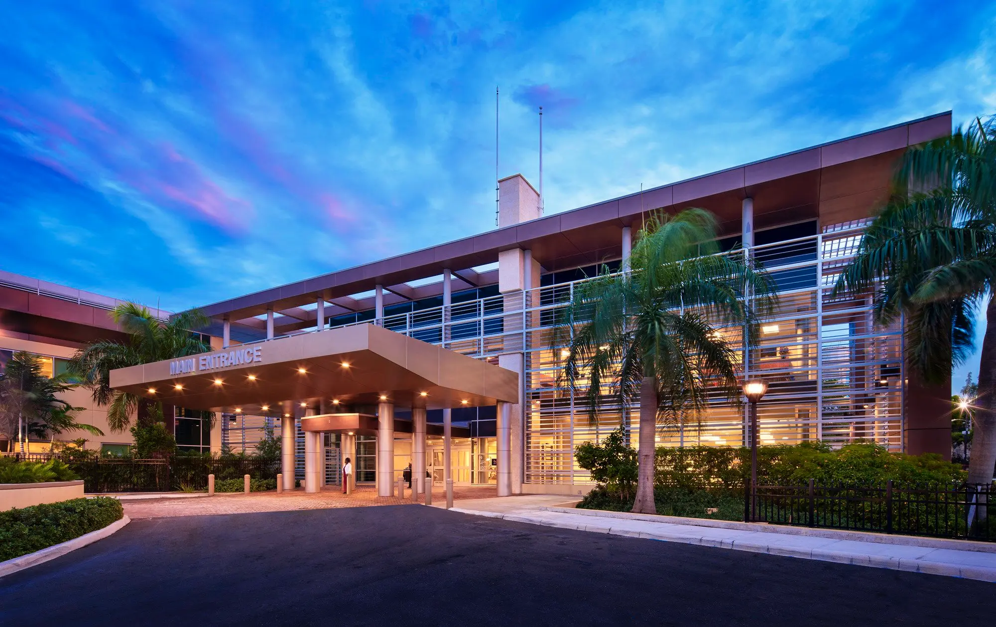 Moderní budova Princess Margaret Hospital v Nassau na Bahamách, obklopená palmami a osvětlená večerním světlem.