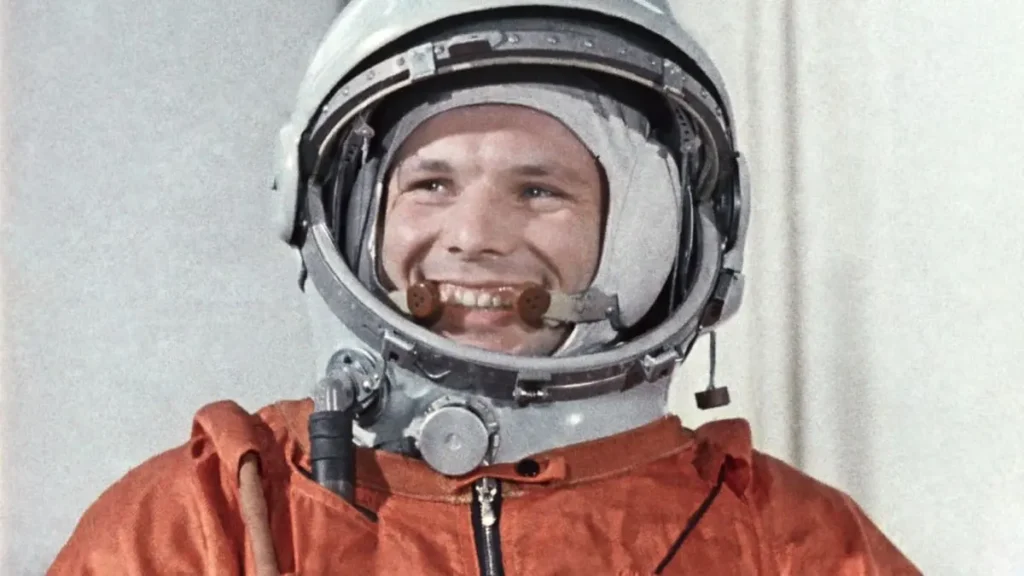 Jurij Gagarin v oranžovém kosmonautském obleku a přilbě, s širokým úsměvem na tváři. Gagarin byl první člověk, který obletěl Zemi ve vesmíru na palubě kosmické lodi Vostok 1 v roce 1961.