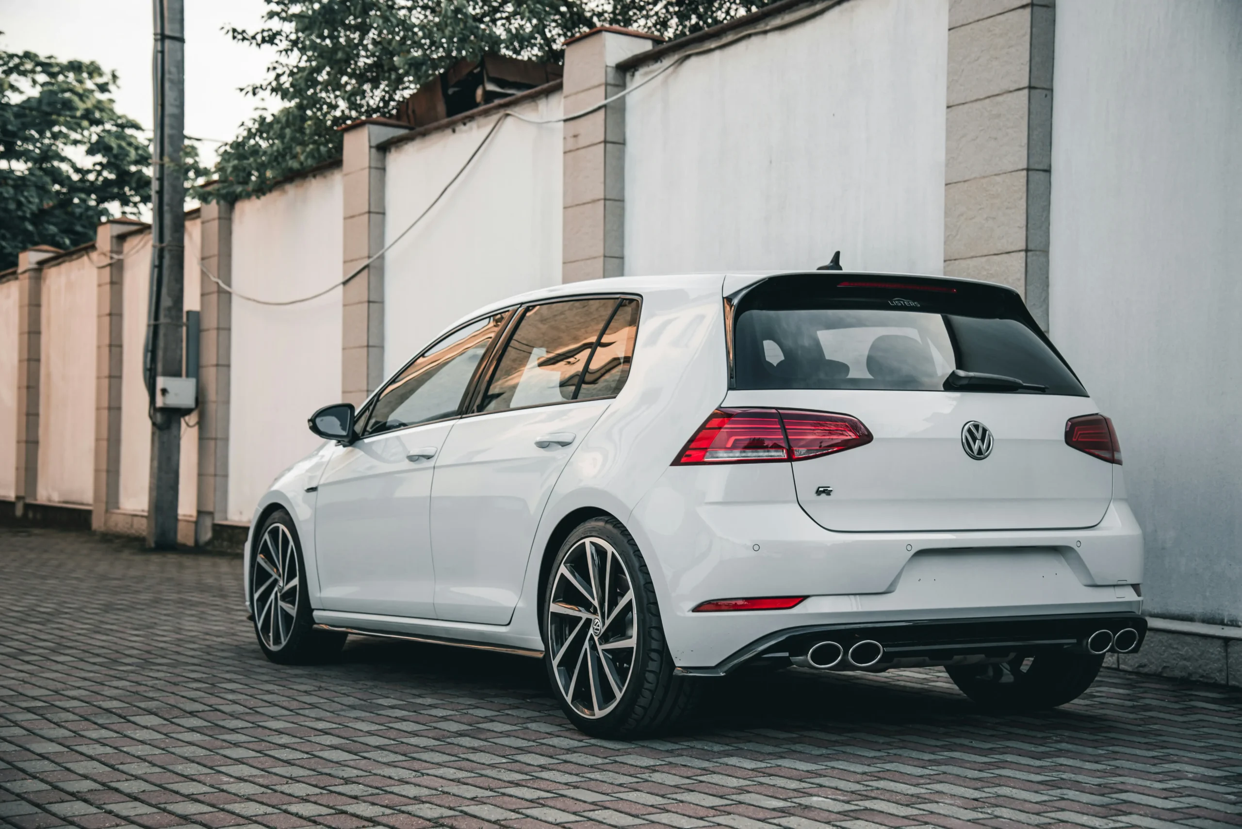 Bílý Volkswagen Golf R s elegantním sportovním designem, čtyřmi koncovkami výfuku a černými detaily zaparkovaný na dlážděné ploše u bílé zdi.