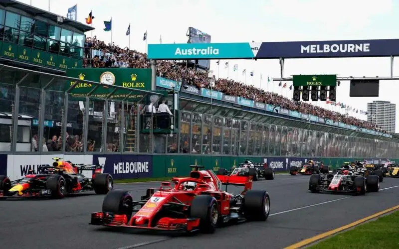 Startovní rošt Velké ceny Austrálie Formule 1 v Melbourne s červeným vozem Ferrari na čele, obklopeným dalšími monoposty, zatímco diváci sledují závod z tribun.