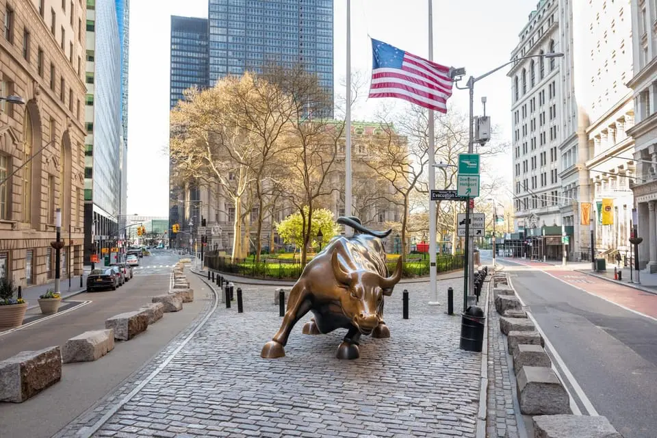 Bronzová socha Charging Bull na Wall Street v New Yorku, symbolizující sílu a optimismus finančních trhů, s americkou vlajkou a městskou architekturou v pozadí.