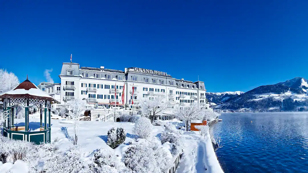 Historický Grand Hotel v Zell am See obklopený zasněženou krajinou a tyrkysovým jezerem pod jasným zimním nebem.