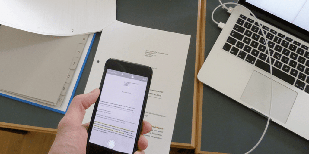 Ruka držící smartphone při skenování dokumentu. Na stole jsou položeny papírové dokumenty, pořadač s oddělovači a otevřený notebook připojený k nabíjecímu kabelu. Proces digitalizace tištěného textu pomocí mobilní aplikace.