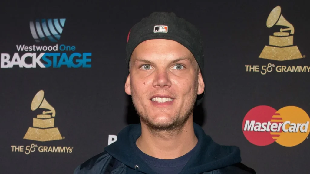 Avicii na červeném koberci před cenami Grammy, oblečený v černé kšiltovce a mikině s logy MasterCard a Grammy v pozadí.