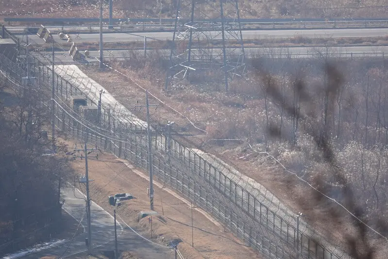 Záběr na ostře střežené demilitarizované pásmo s ploty, strážními věžemi a vojenskými prvky na hranici mezi Severní a Jižní Koreou. Scéna zachycuje infrastrukturu a drsné podmínky této oblasti.