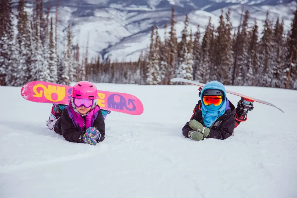 Dvě děti odpočívající na sněhu s barevnými snowboardy, oblečené v zimním oblečení, připravené na další jízdu.